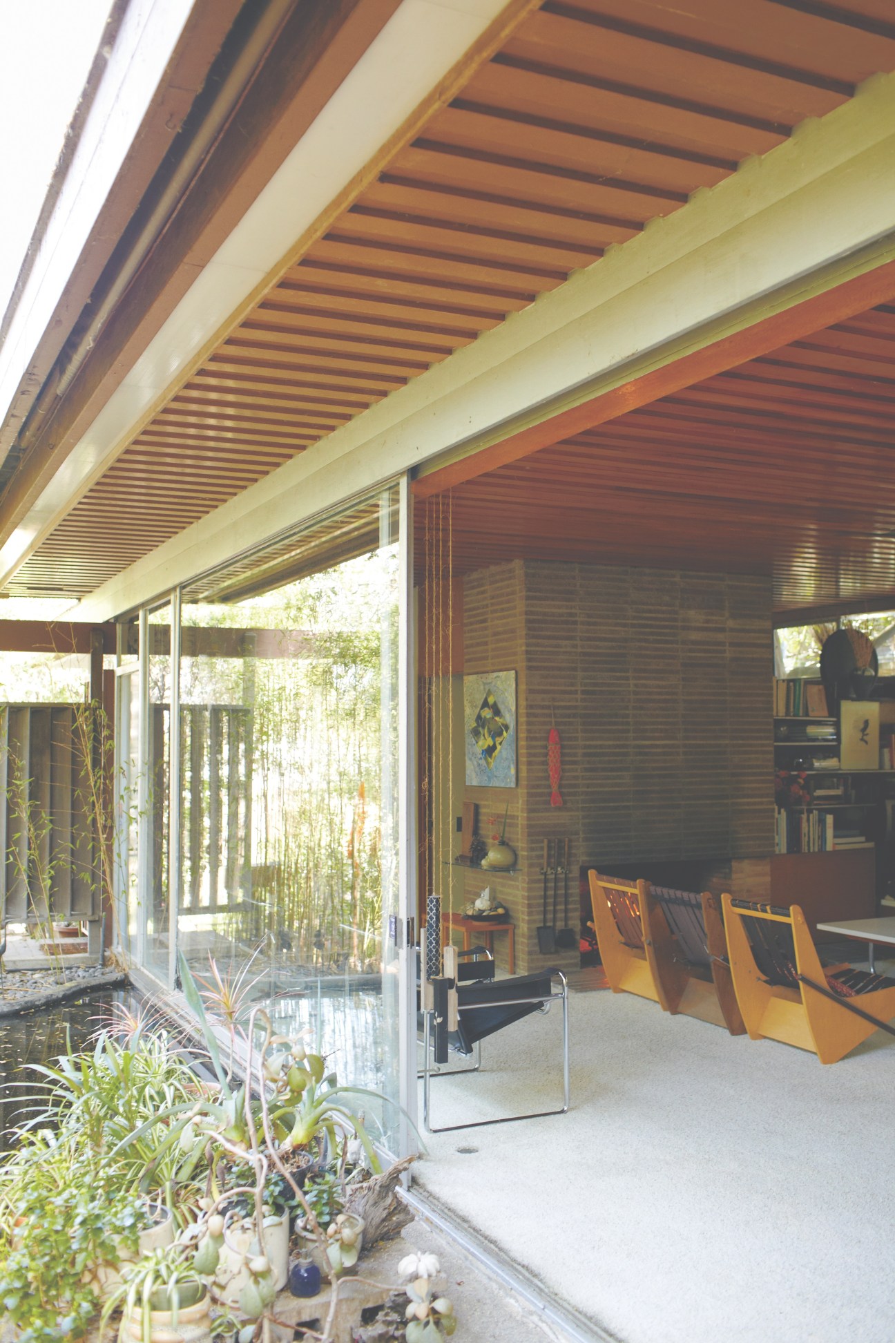 Glass sliding doors open from a modernist interior with wood paneling and mid-century chairs to a garden with potted plants.
