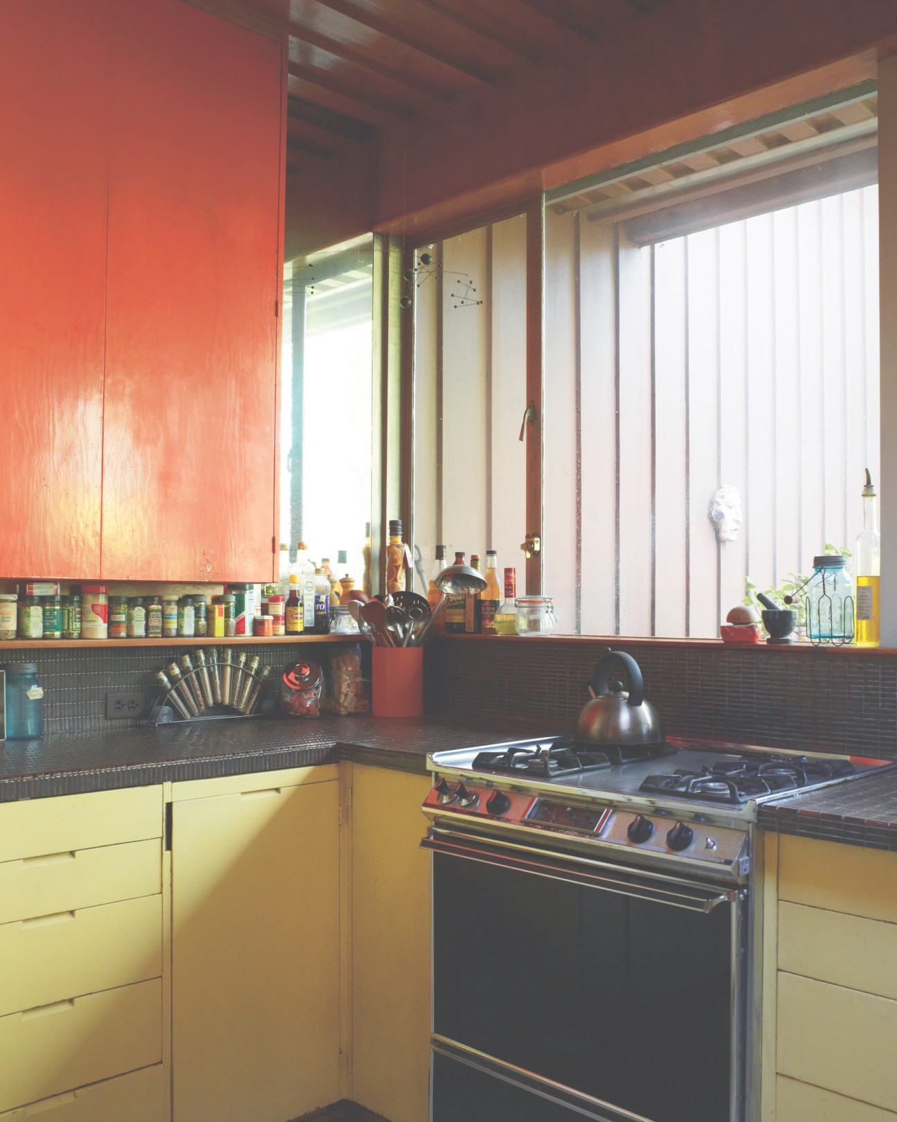 Mid-century modern kitchen with yellow cabinets, red overhead panel, black countertop, and large windows overlooking the garden.