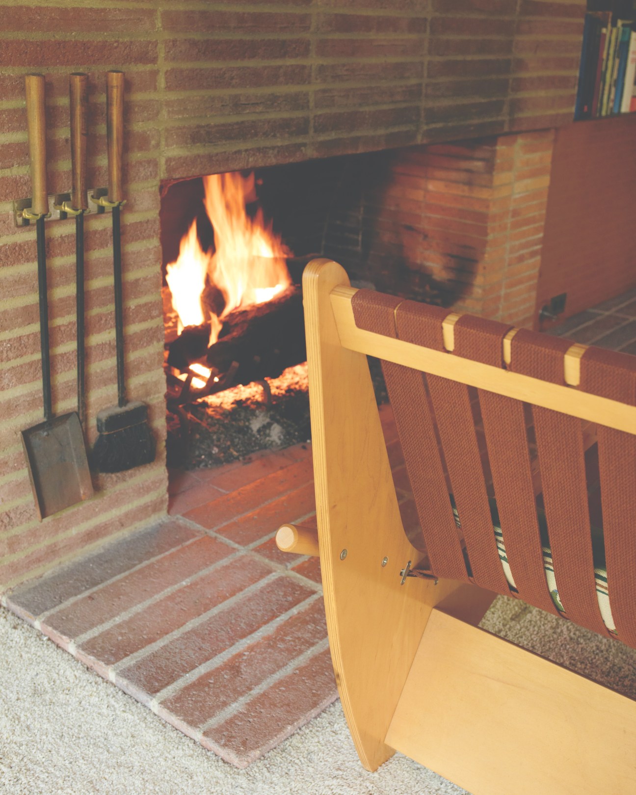 A Wassily chair with red webbing sits beside a brick fireplace with active flames and metal tools.