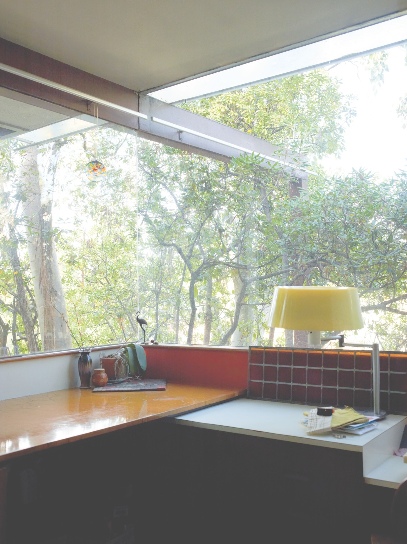 Master bedroom with desk overlooking lush garden through floor-to-ceiling glass walls, featuring a yellow lamp and spider-leg roof support.