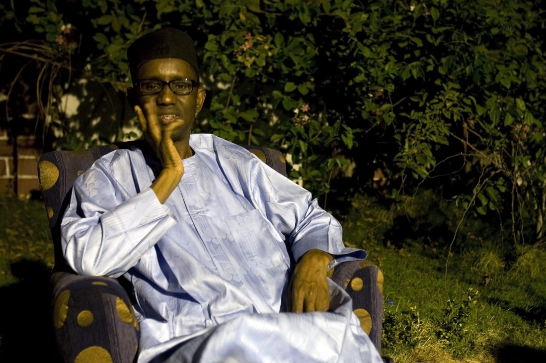Nuhu Ribadu sits on a chair outdoors, wearing a light blue shirt and glasses, hand to his face in a contemplative pose.