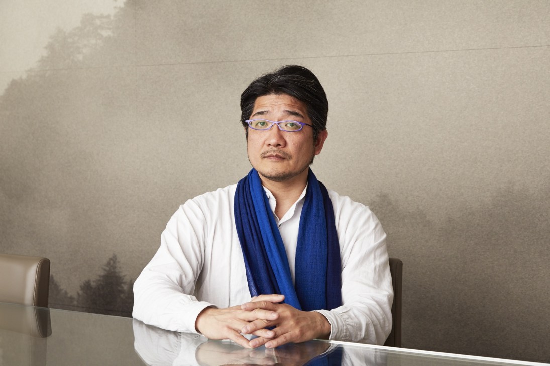 Portrait of Hiroshi Seki, a man wearing glasses, white shirt and blue scarf, seated at a table.