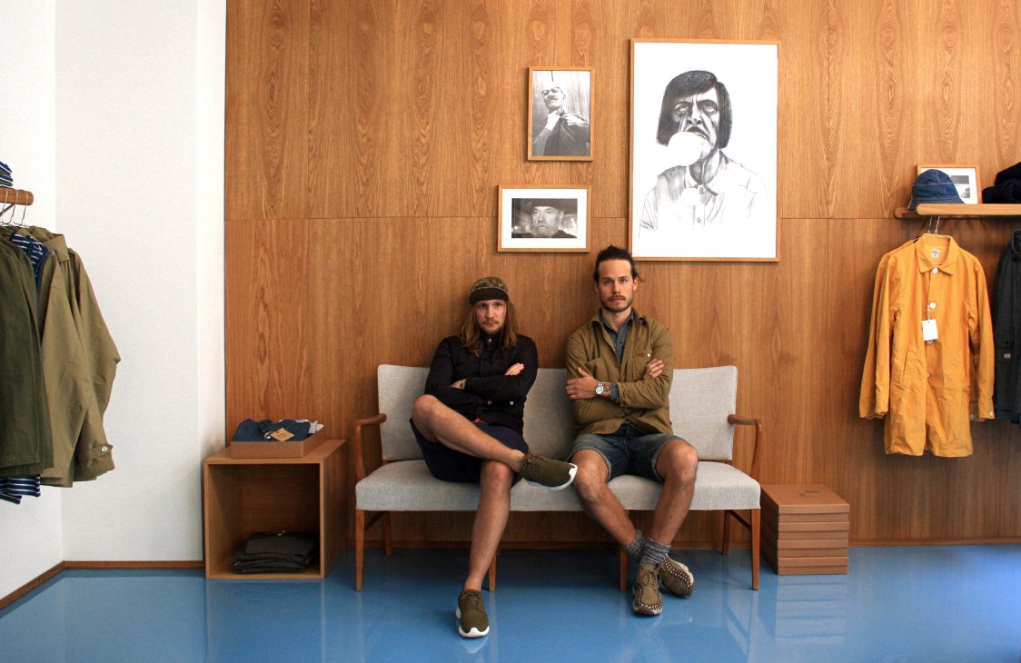 Two men sit on a grey sofa against a wood-panelled wall with framed portraits, wearing casual clothing in a retail store setting.