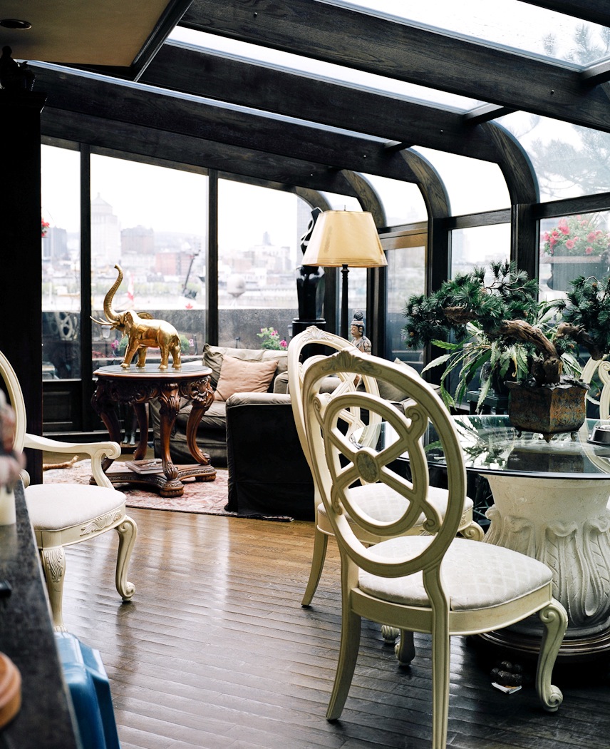 Enclosed terrace with green ornate chairs, gold elephant statue, and cityscape view through large windows.