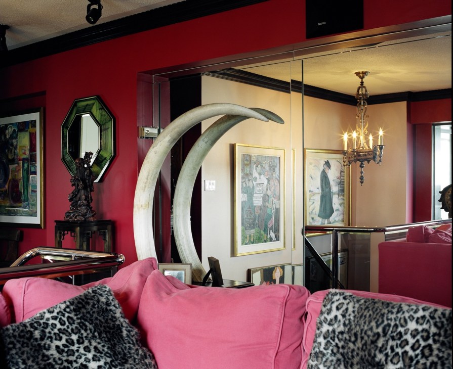 A sitting room with vibrant red walls, pink sofa with leopard print pillows, large curved ivory tusks, framed artwork, and brass chandelier.