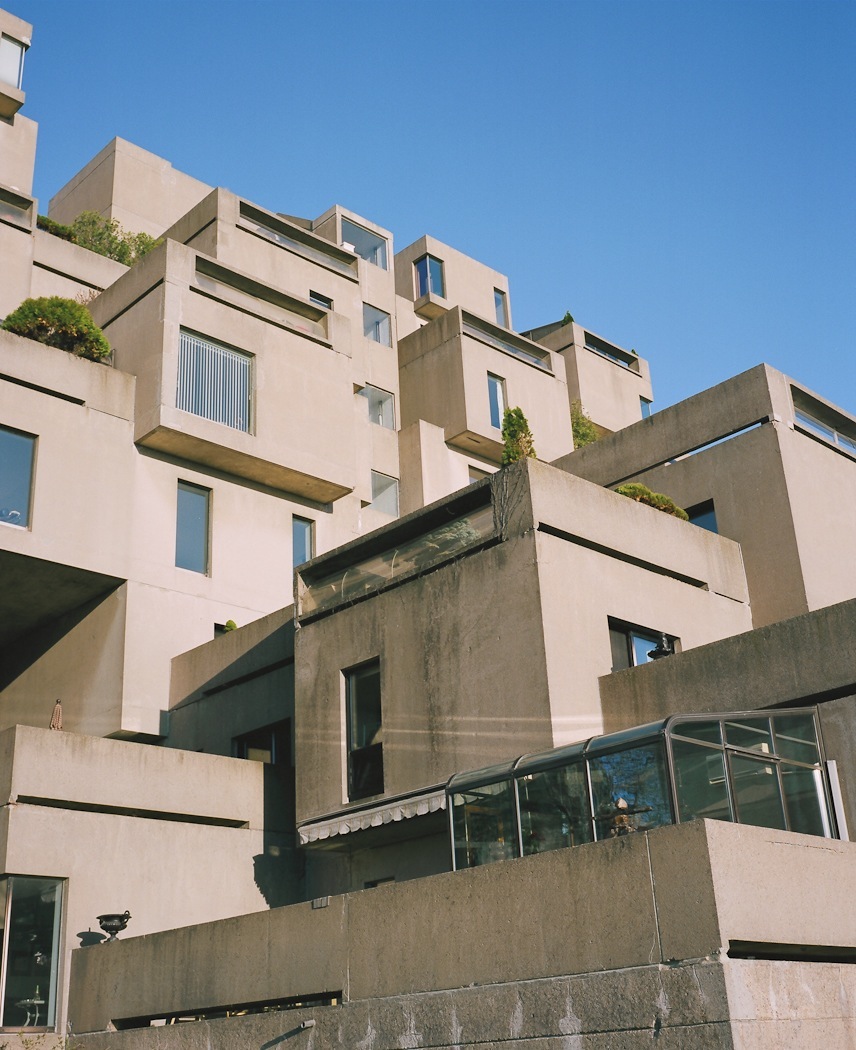 Stacked concrete cube units of Habitat 67 with staggered terraces and vegetation against blue sky.