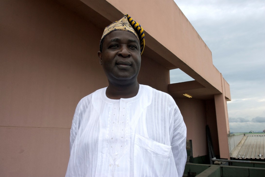 Diran Kolajo, a civil servant in white traditional attire and yellow headwrap, stands against a modern terracotta building.