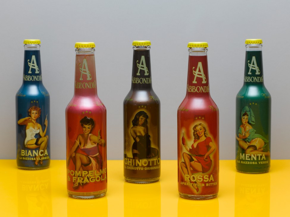 Five Abbondio vintage-style glass bottles with yellow caps and illustrated pin-up labels on a yellow surface.