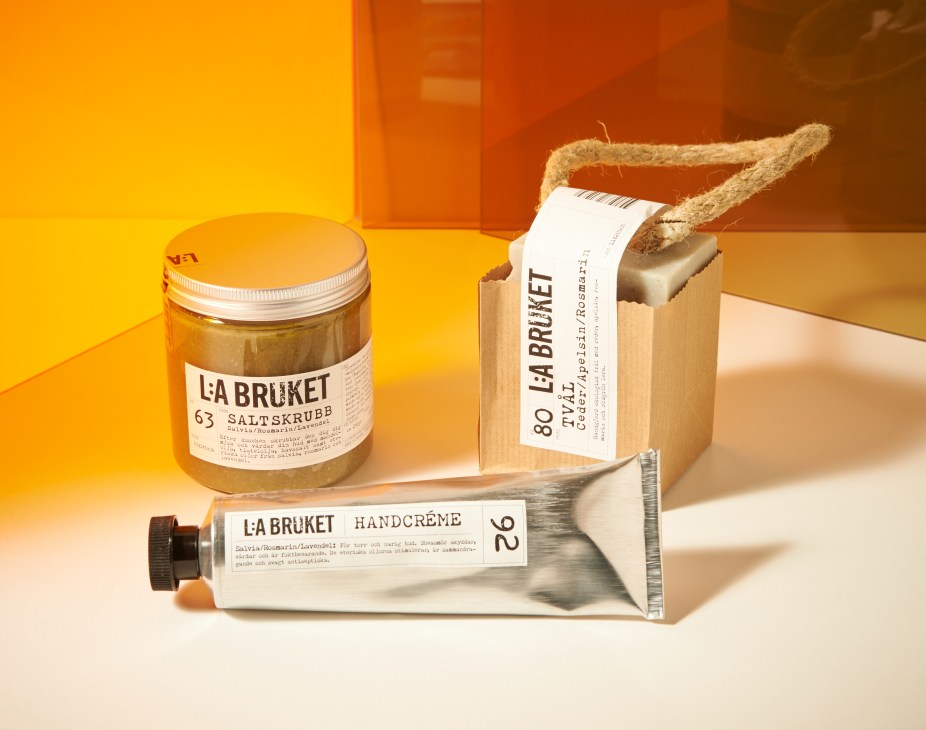 L:A Bruket skincare products including a salt scrub jar, hand cream tube, and soap bar with rope handle against a warm orange background.