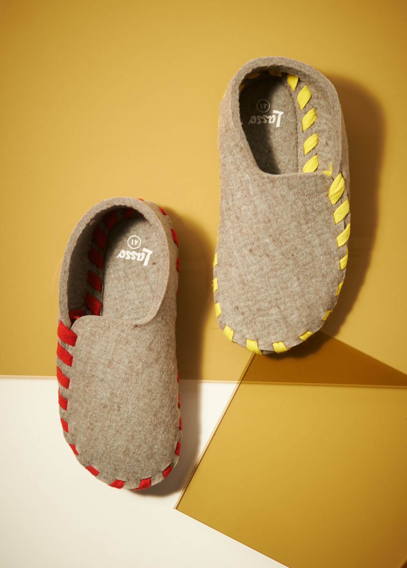 Two Lasso felt slippers with colored laces in red and yellow against a yellow background.