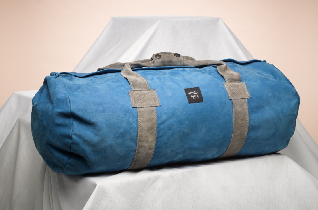 Blue Beastin leather duffle bag with grey suede straps and metal handles on white fabric.