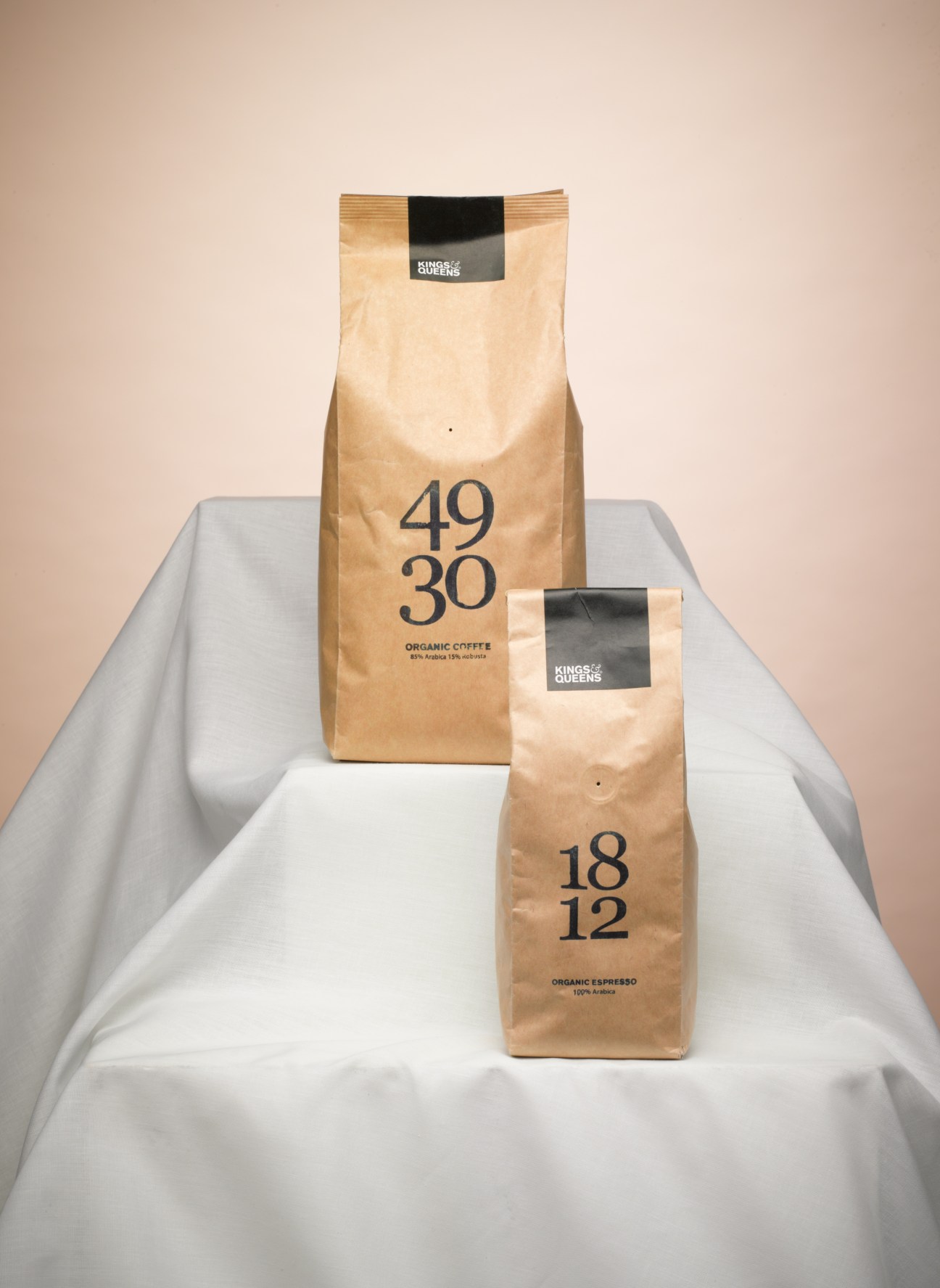 Two kraft paper bags of Kings&Queens organic coffee with black labels on a white draped fabric against a beige background.