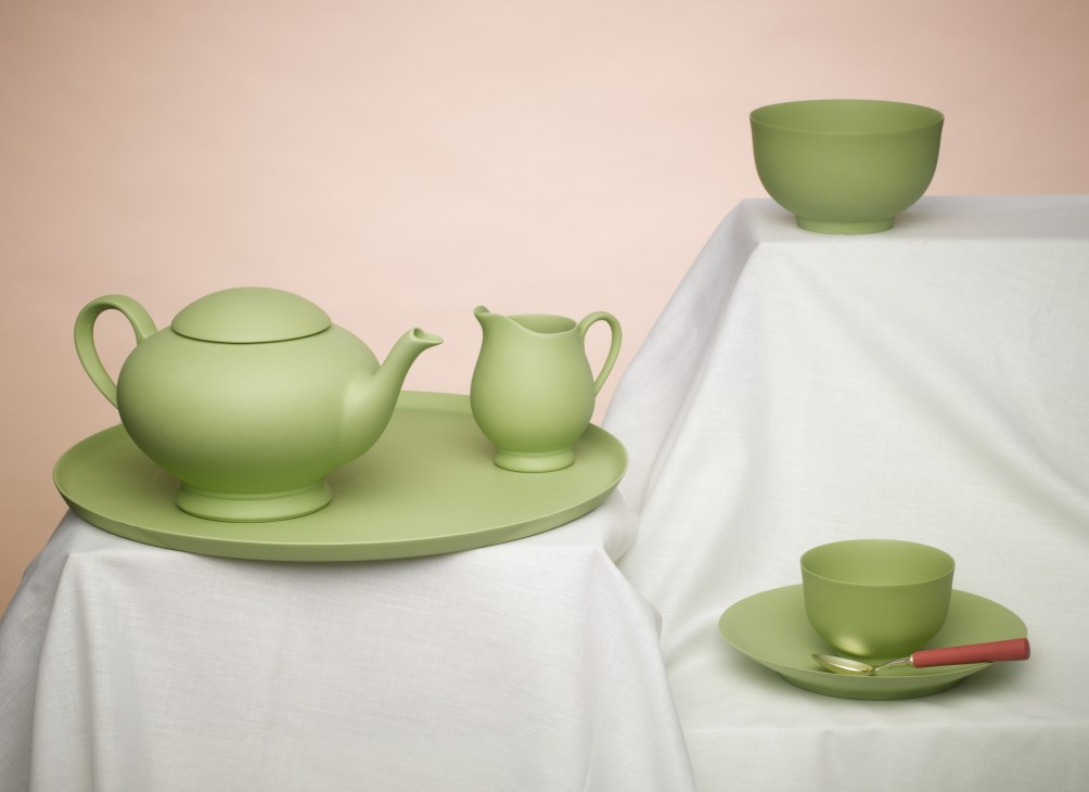 Green ceramic tableware set including teapot, pitcher, bowls and cup on white cloth.