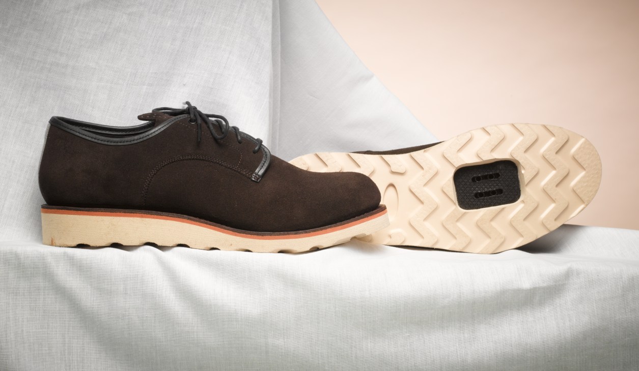 Brown suede shoe with cream and orange sole displayed on white fabric.