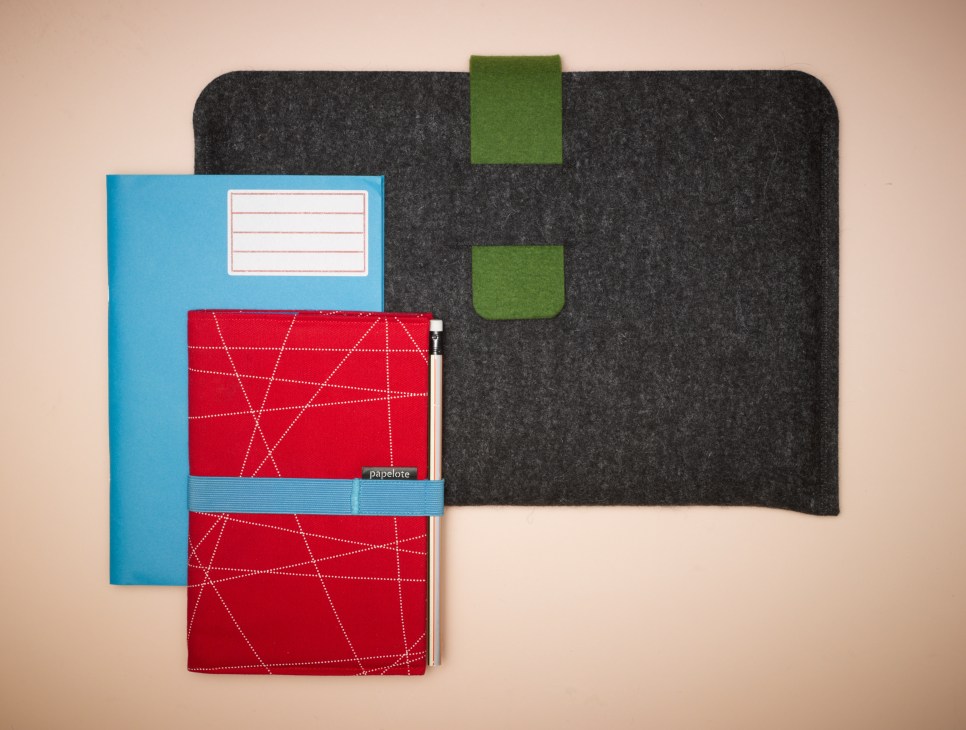 Papelote stationery collection featuring blue notebook, red geometric patterned folders, and charcoal felt laptop sleeve with green accents.