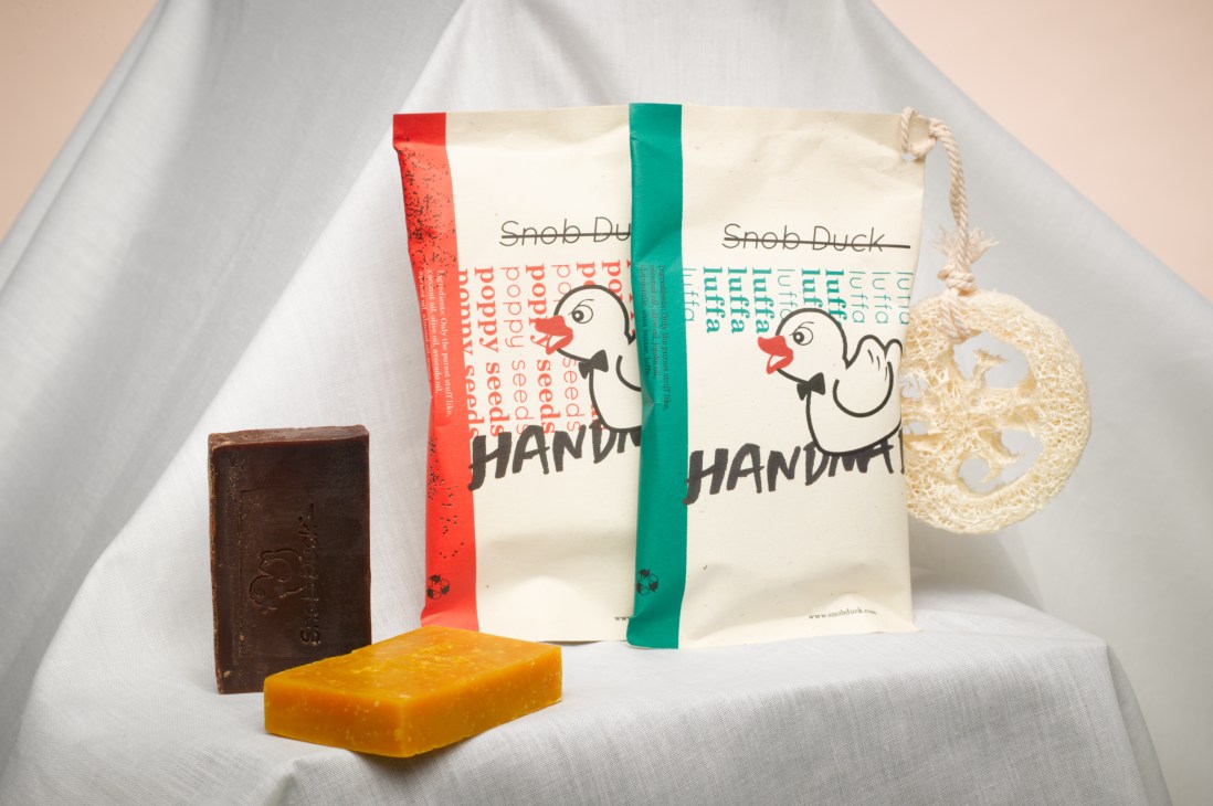 Snob Duck handmade soaps in red and green packaging with natural olive oil bars displayed on white fabric.