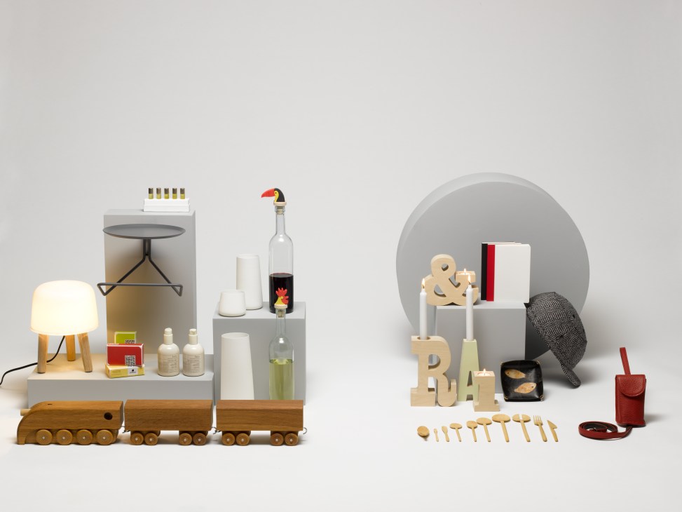 Styled product arrangement featuring a wooden toy train, lamp, nail oils, bottle stopper, cutlery, candle holders, notebook, leather bowl, and bike helmet accessories.