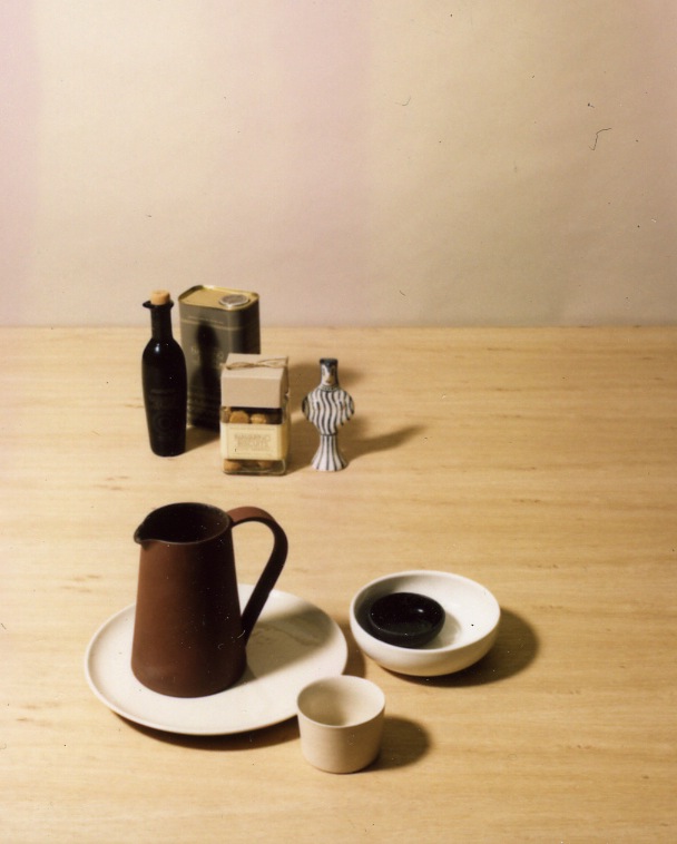 Another Country tableware collection featuring clay jug, bowls, plate and cup in various earth tones arranged on wooden surface.