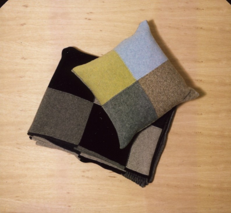 Esk Valley Knitwear cushion and throw in grey, black, yellow and blue tones on wooden floor.