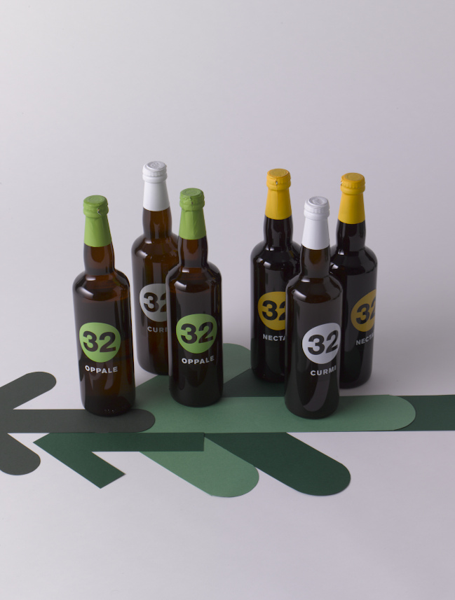 Six beer bottles from 32 Via dei Birrai arranged on a green decorative base against a grey background.