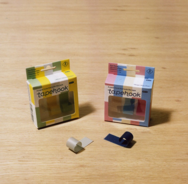 Torafu Architects Tapehook packaging in yellow and multicolored boxes with white and navy hook samples on wooden surface.
