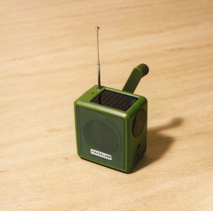 Green compact radio with solar panel on top, hand-crank handle, and extendable antenna on wooden surface.