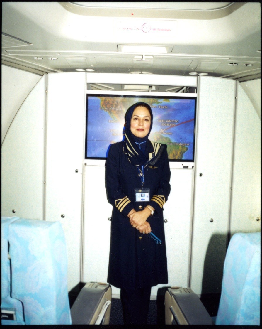 IranAir stewardess in dark blue uniform with gold trim and headscarf standing in aircraft cabin.