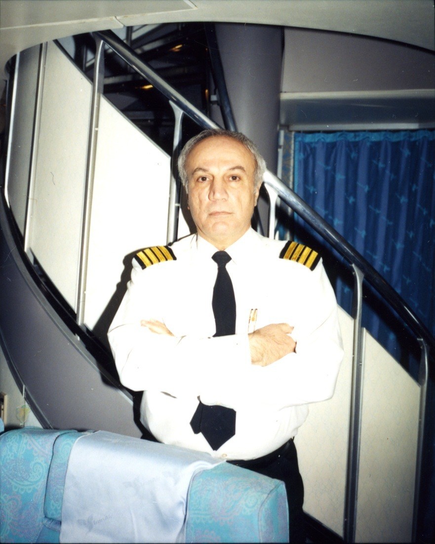 Captain James Farrahi in white uniform with gold-striped shoulders and black tie sitting in aircraft cabin.