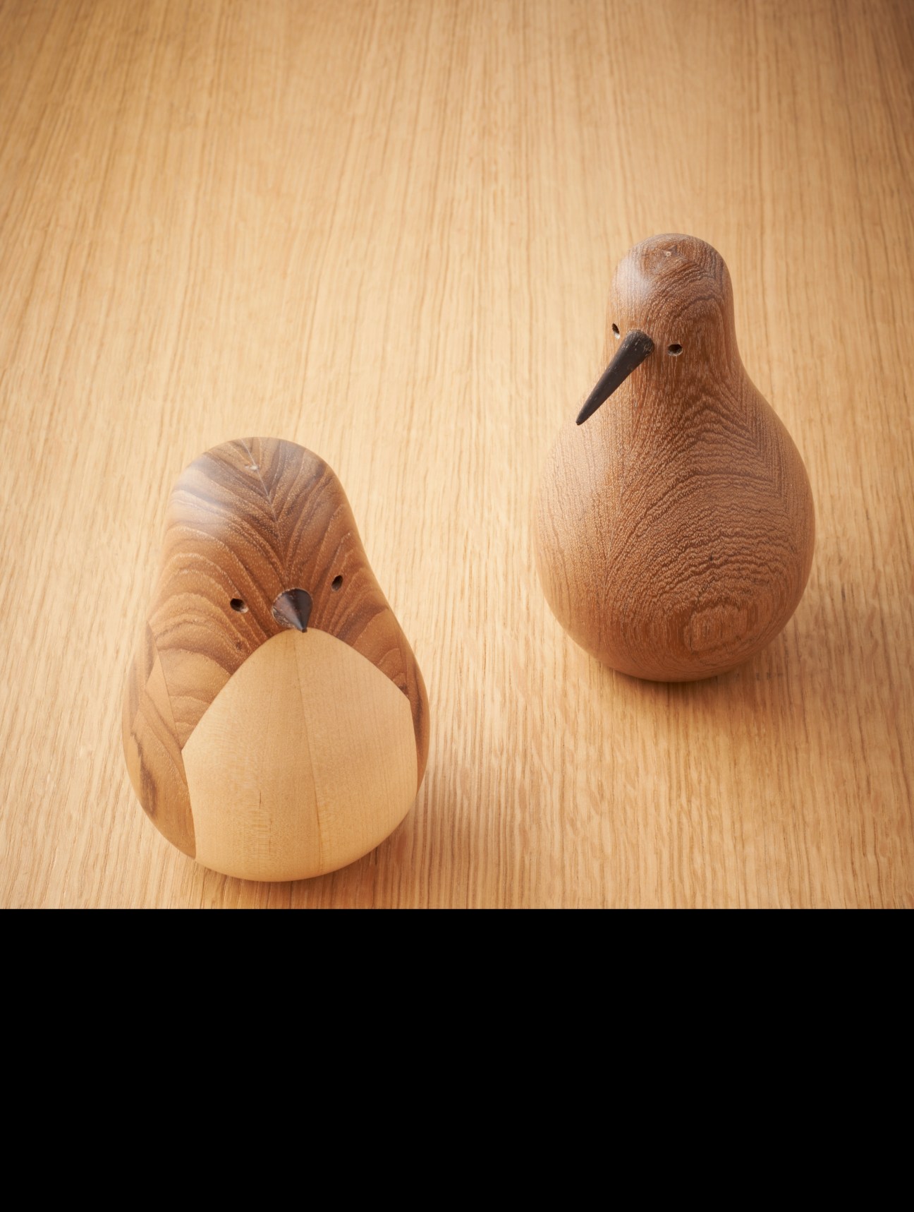 Two wooden house bird sculptures in natural wood tones on a light wood surface by Lars Beller Fjetland.