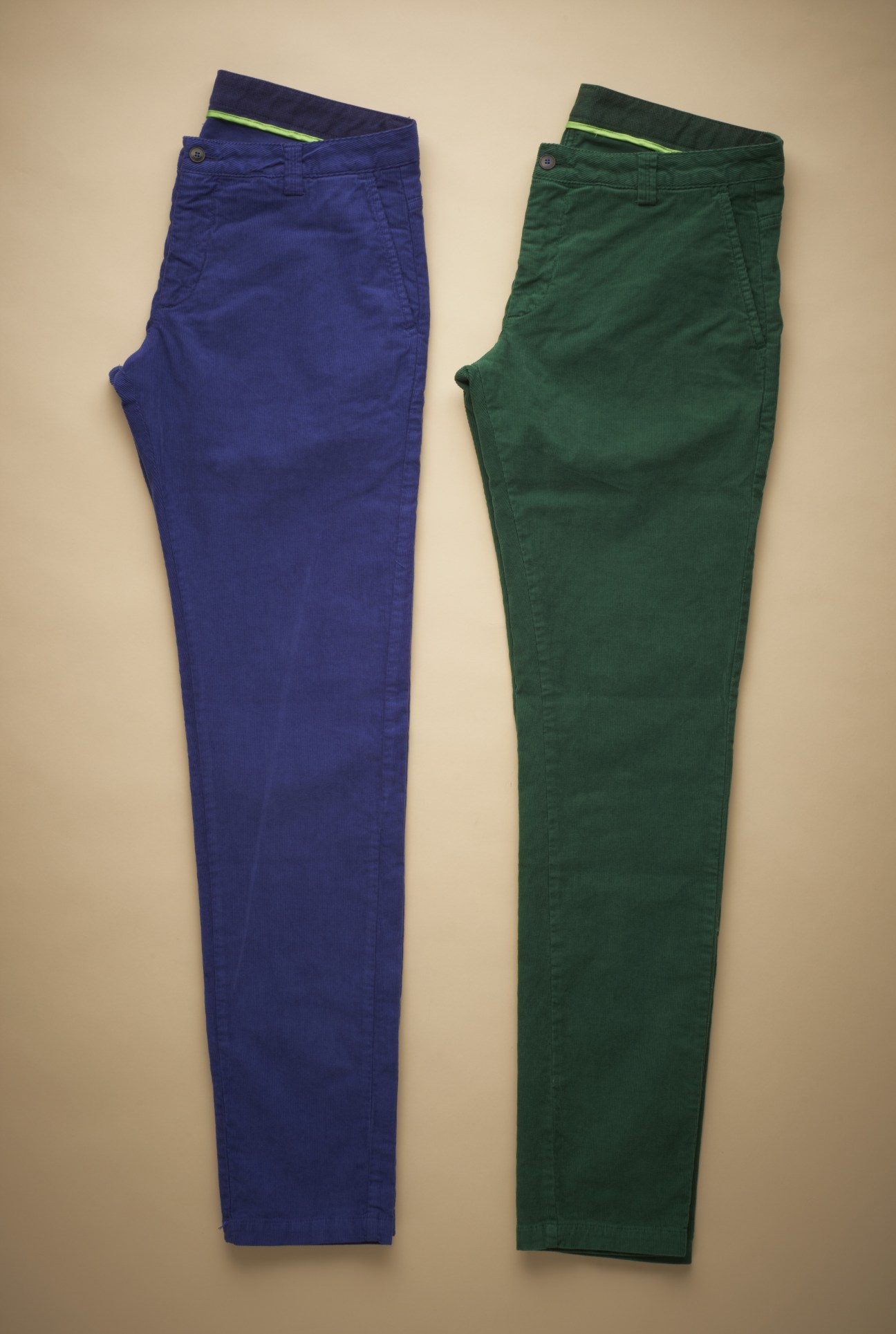 Two pairs of flat-lay trousers in navy blue and forest green with neon yellow waistband details against a beige background.