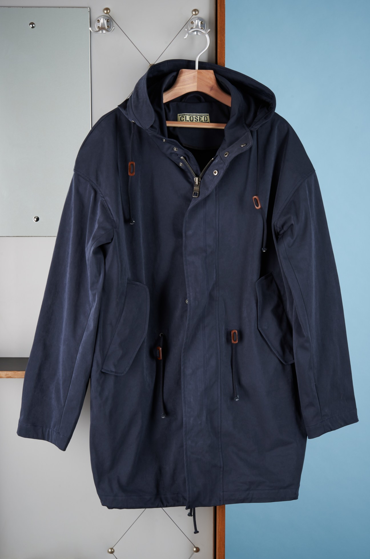 Navy hooded chino jacket by Closed displayed on a wooden hanger against a white and blue wall.