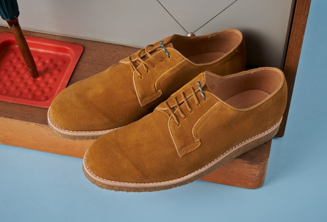 Pair of tan suede desert boots with crepe soles displayed on wooden blocks against a blue background.