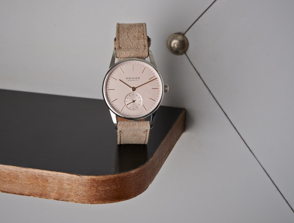 Nomos Orion 33 rosé wristwatch with rose gold dial, stainless steel case, and beige velour strap on wooden display.