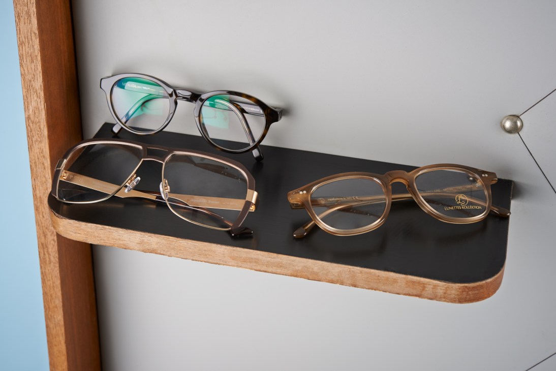 Three pairs of eyeglasses displayed on a wooden and black shelf against a light background.
