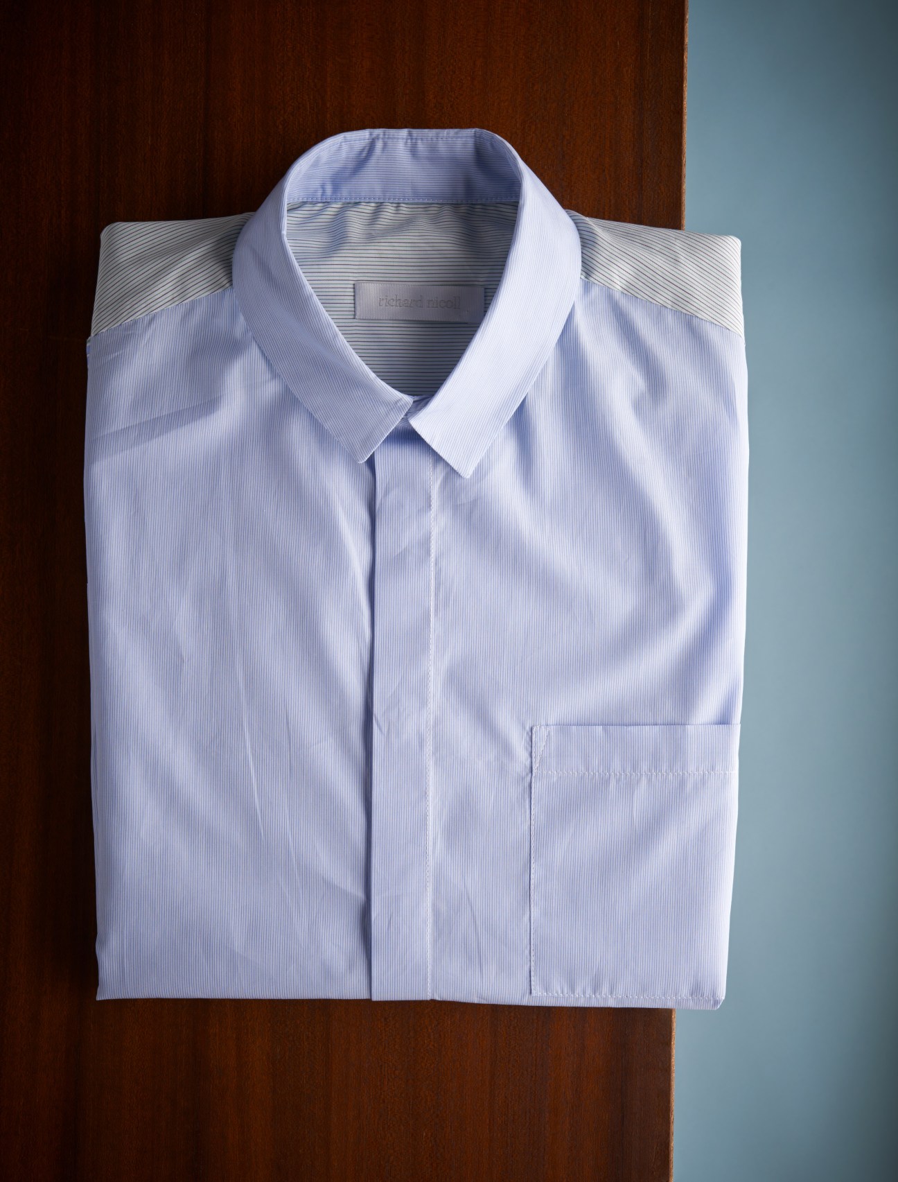 Folded light blue striped cotton shirt with white collar displayed on brown and blue background.