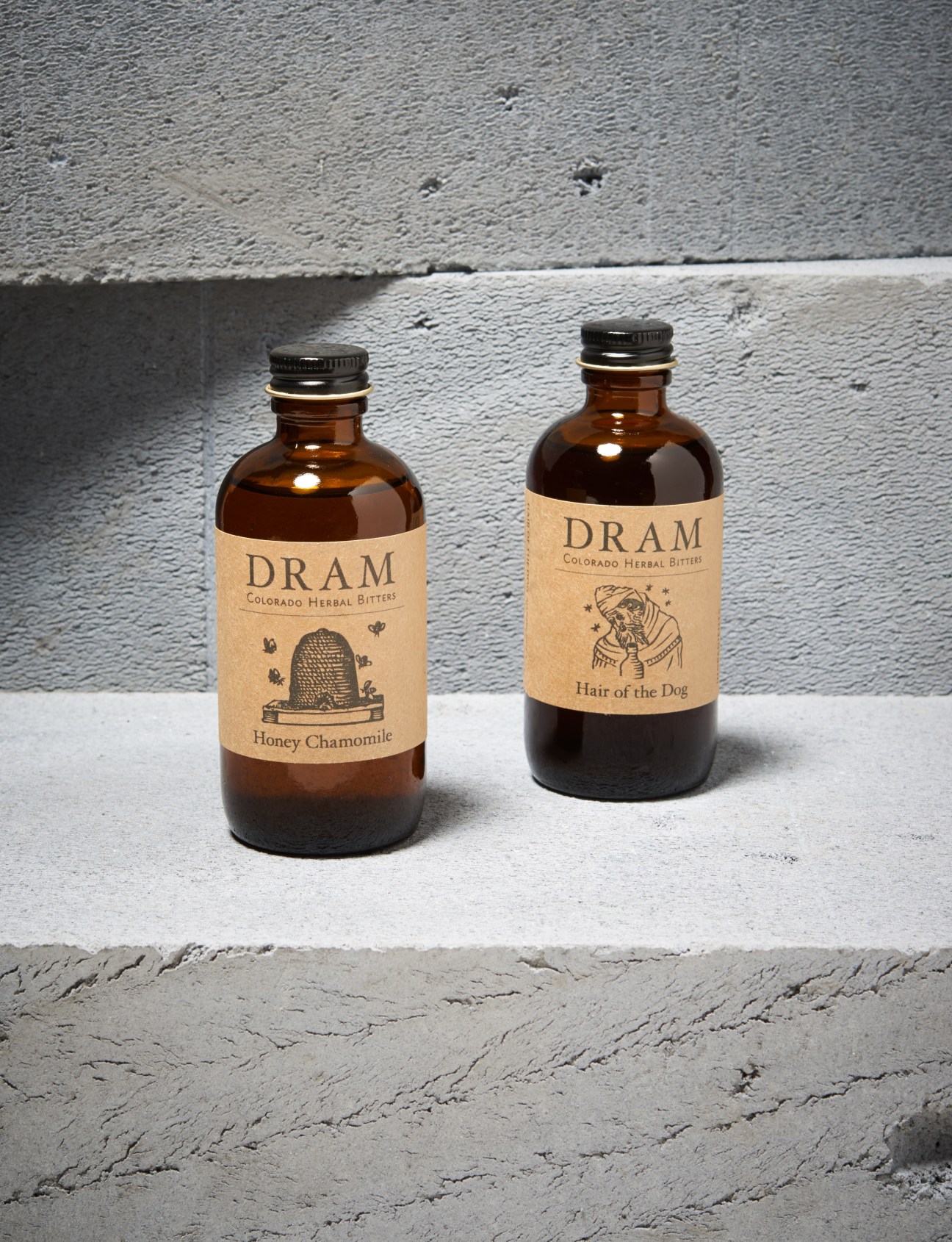 Two amber glass bottles of Dram Colorado Herbal Bitters with kraft labels on a concrete ledge.