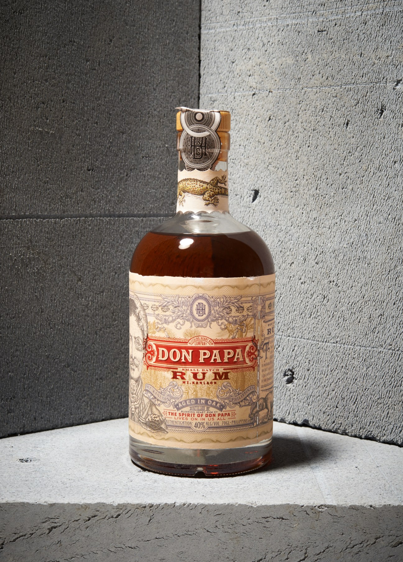 A bottle of Don Papa rum with ornate cream and gold label against a concrete wall backdrop.