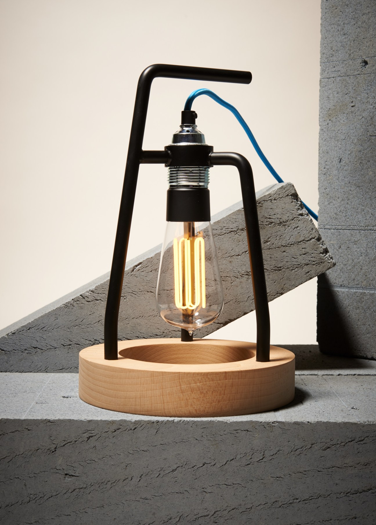 Ministry of Mass barrel lamp with black metal frame, wooden base, and illuminated filament bulb on concrete surface.
