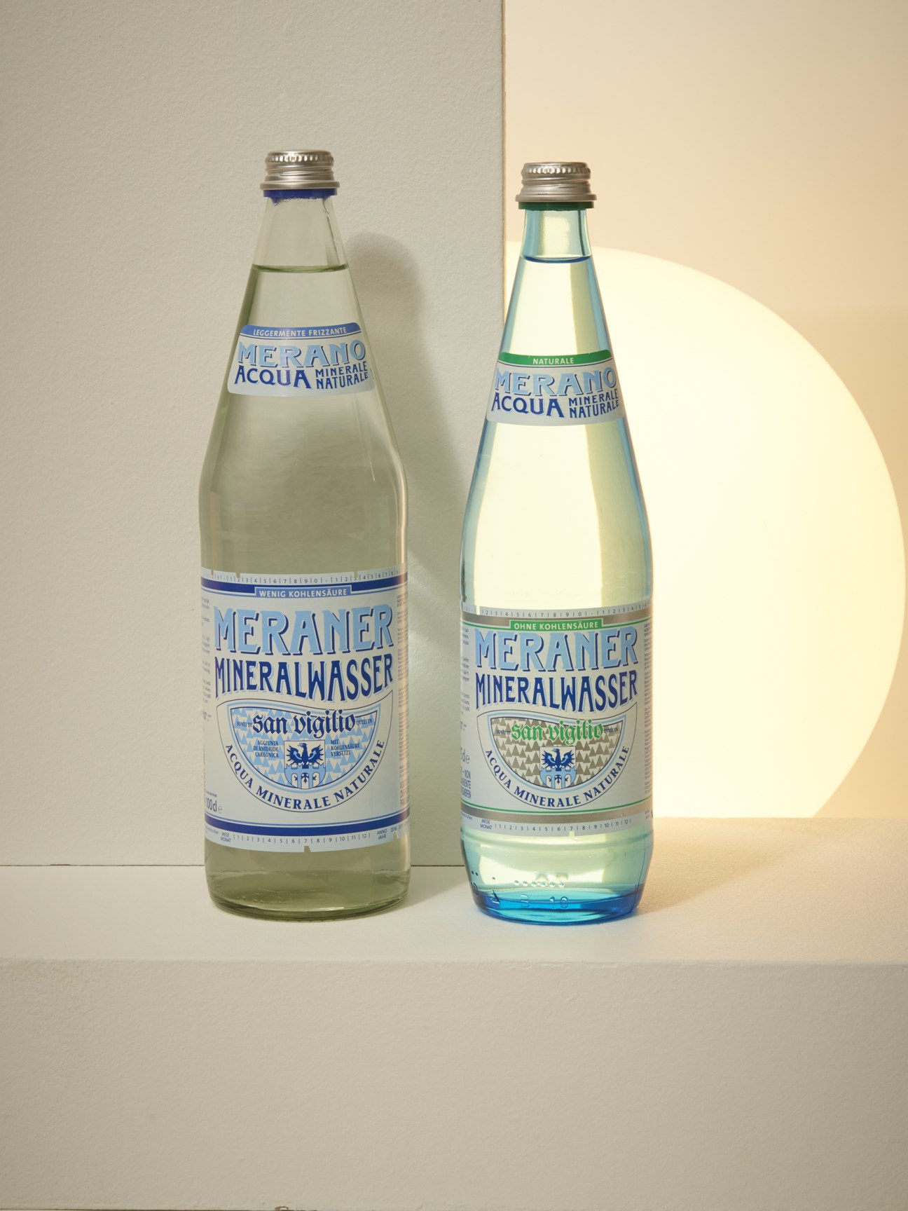 Two bottles of Meraner Mineralwasser mineral water with blue and green labels against a cream-colored background.