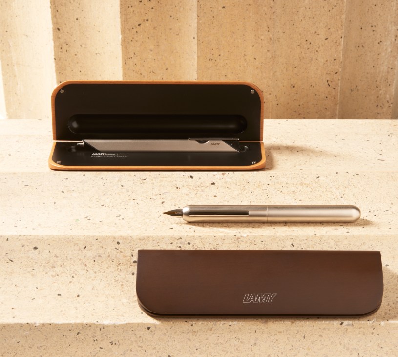 Lamy Dialog series pens in titanium and palladium finishes with brown leather cases on concrete surface.