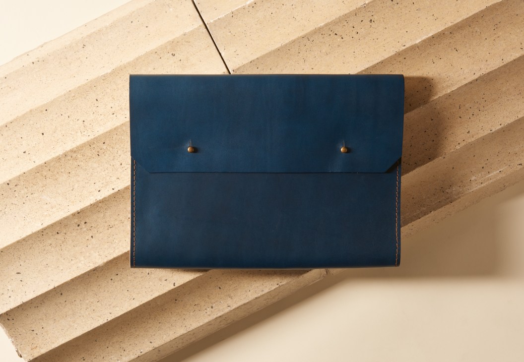 Indigo vegetable-tanned leather document wallet by Noise Goods with brass fasteners on concrete surface.