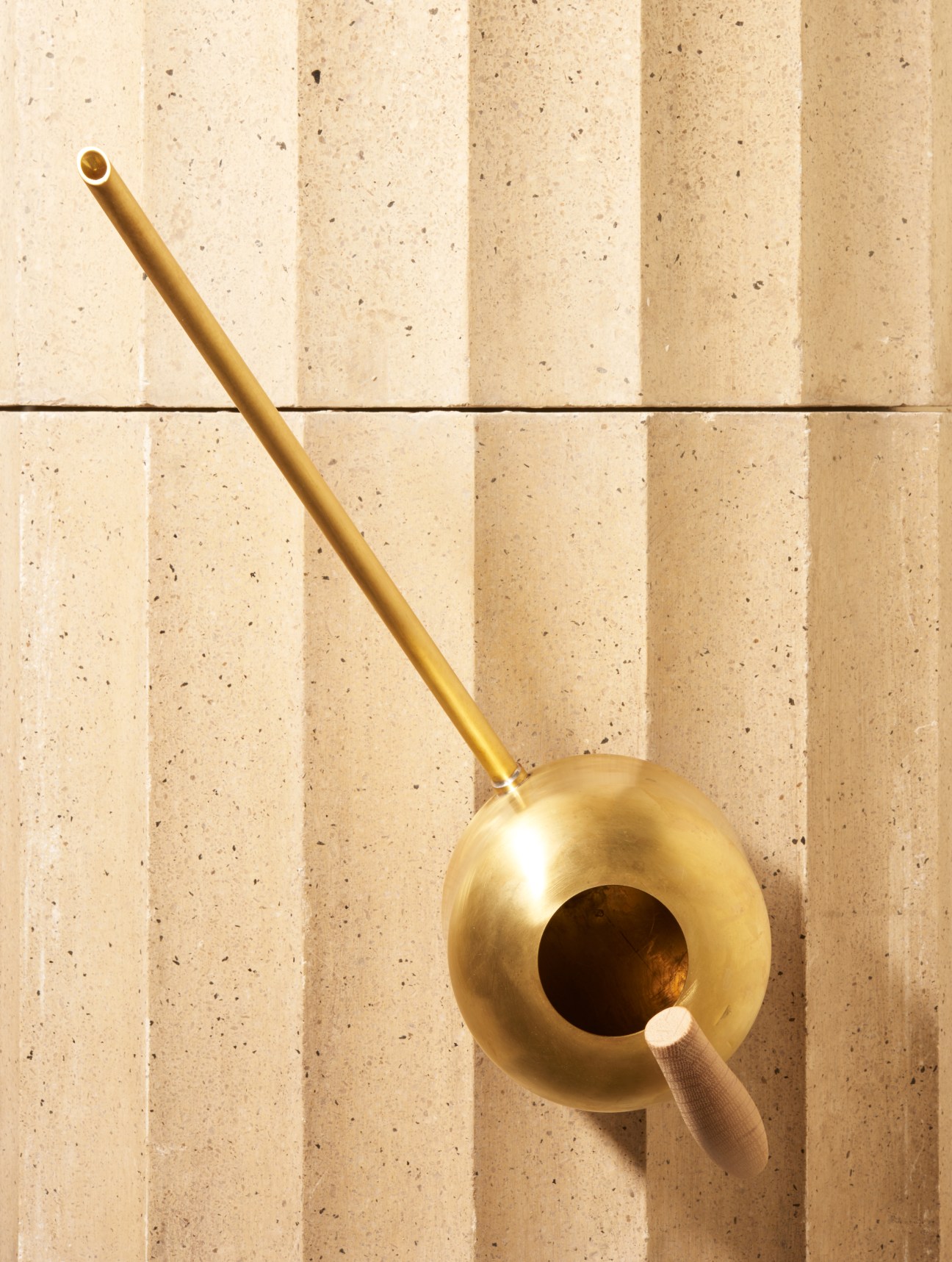Brass watering can with wooden handle against textured beige wall by Mjölk and Anderssen Voll.