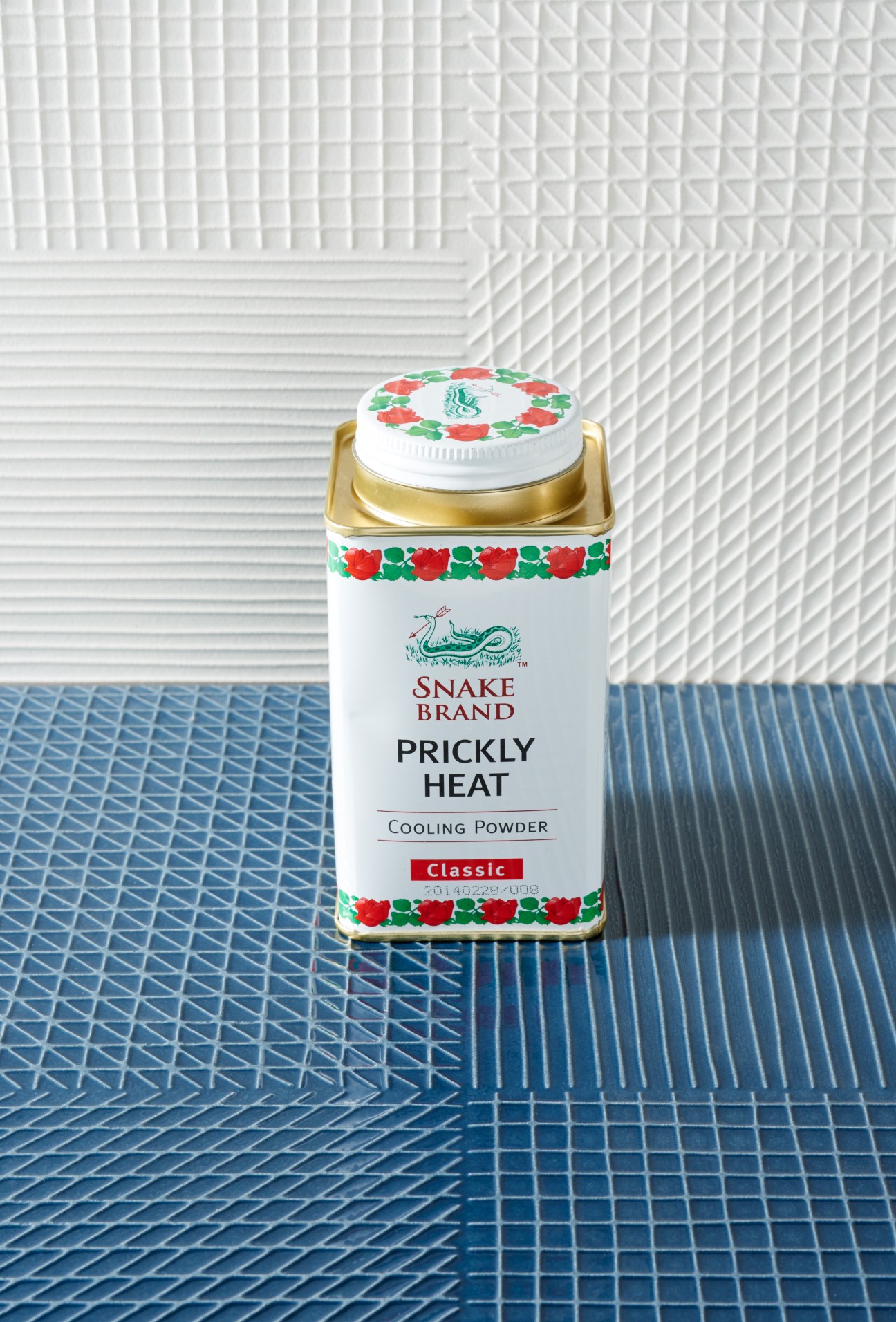 Snake Brand prickly heat cooling powder in white tin with red and green floral decoration against geometric tile backdrop.
