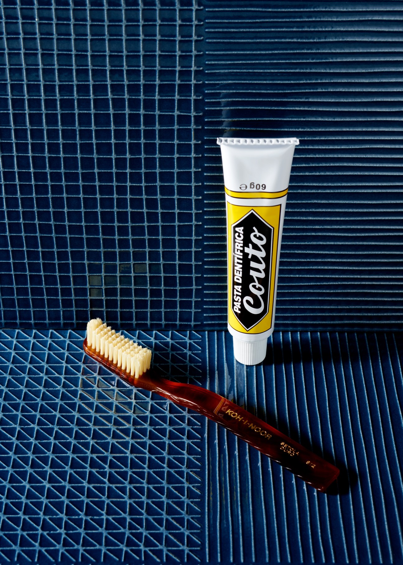 Koh-I-Noor toothbrush with brown handle next to Couto minty toothpaste tube on blue textured tiles.