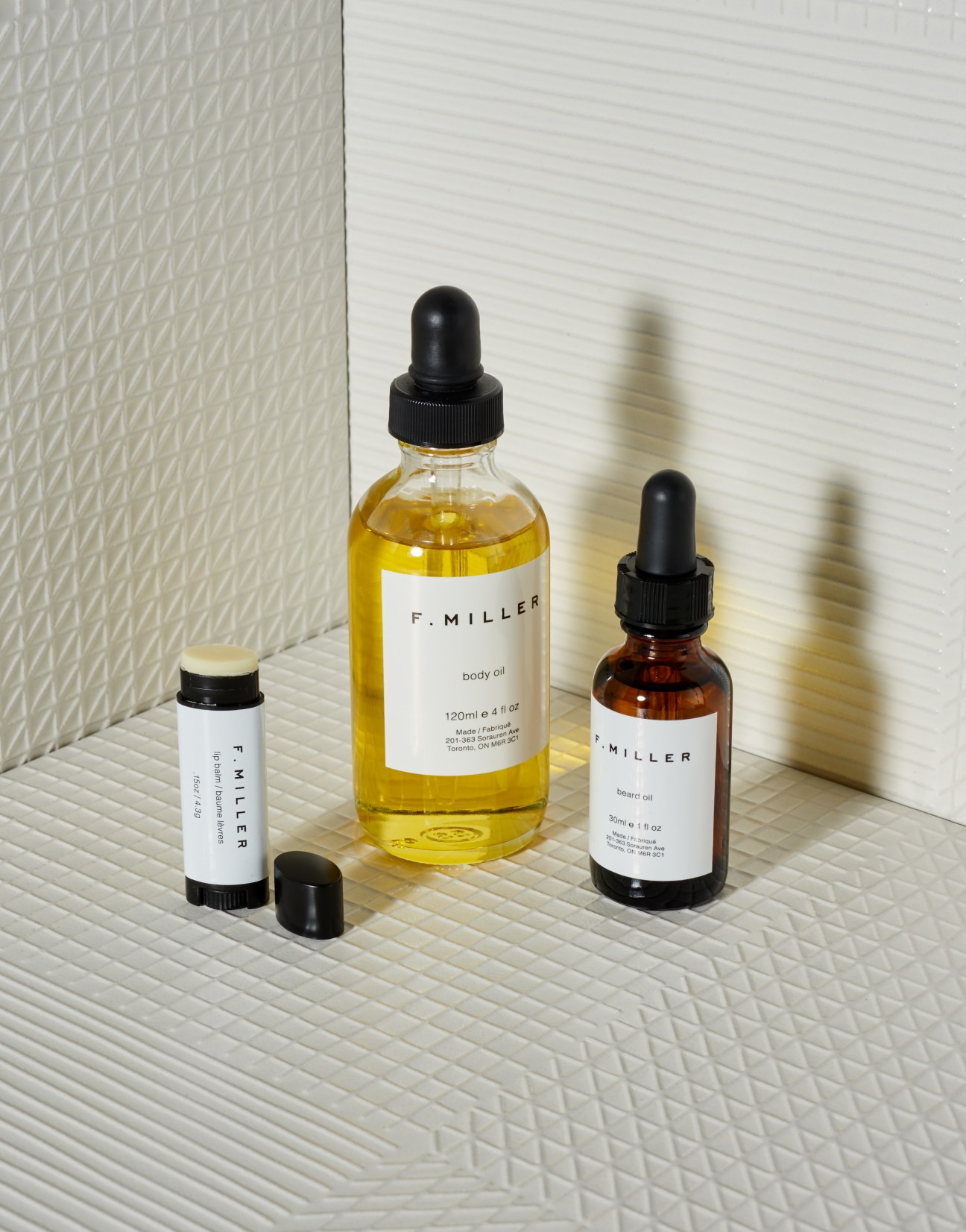 Three F Miller skincare products: lip balm, yellow body oil, and amber facial oil on white textured surface.