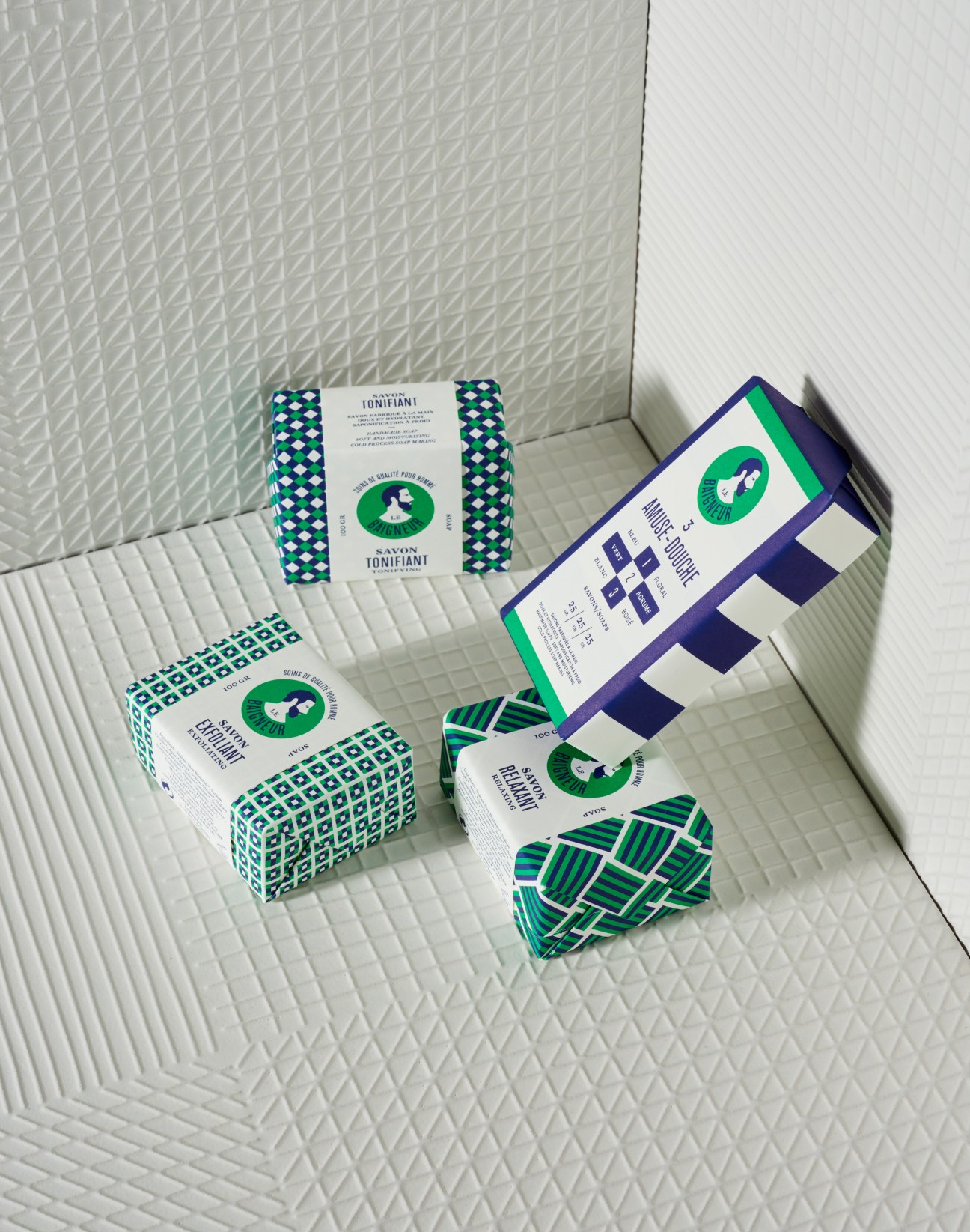 Le Baigneur natural soap bars with green patterned packaging arranged on textured white tiles.