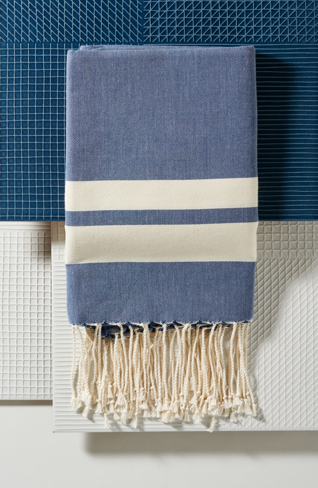 Blue and cream striped cotton towel with fringe ends displayed on patterned tile background.