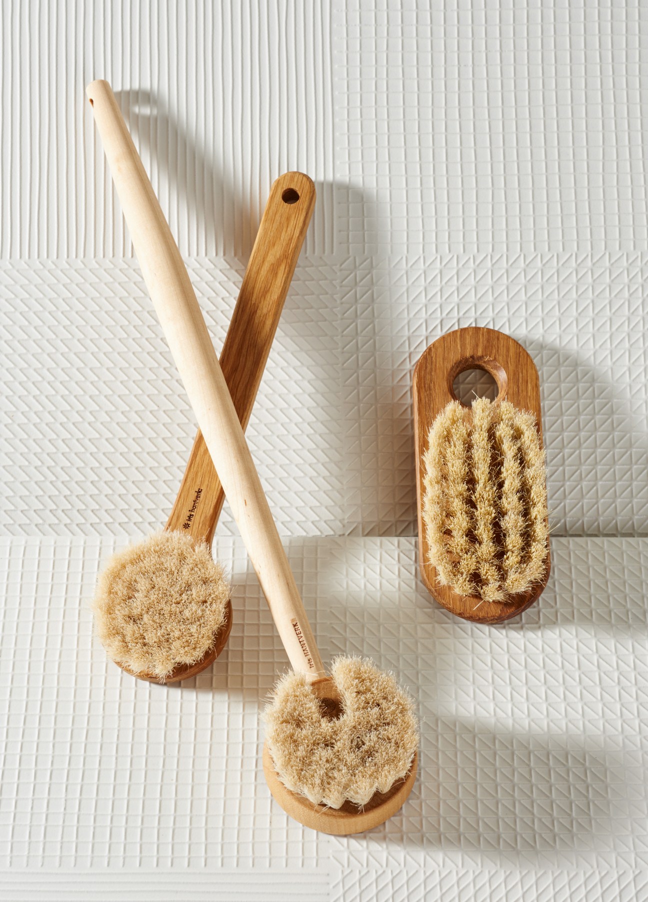 Three wooden-handled body brushes with natural bristles by Iris Hantverk arranged on a textured white tile background.