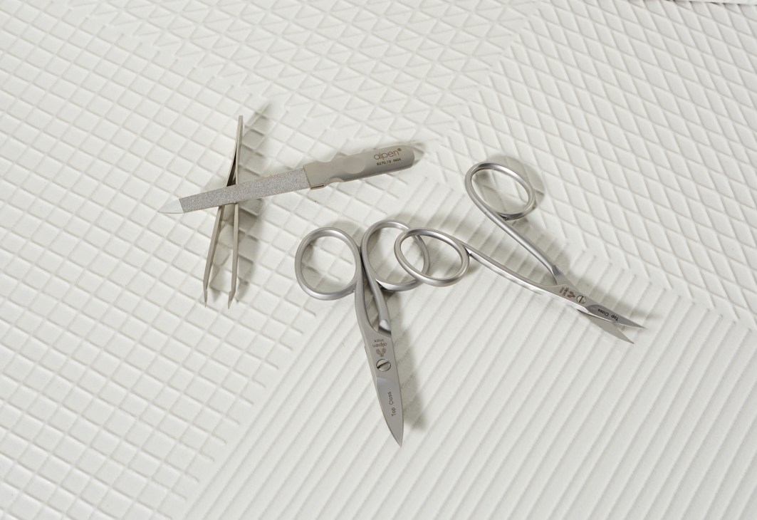 Manicure set by Alpen with nail file, tweezers, and scissors arranged on white textured background.
