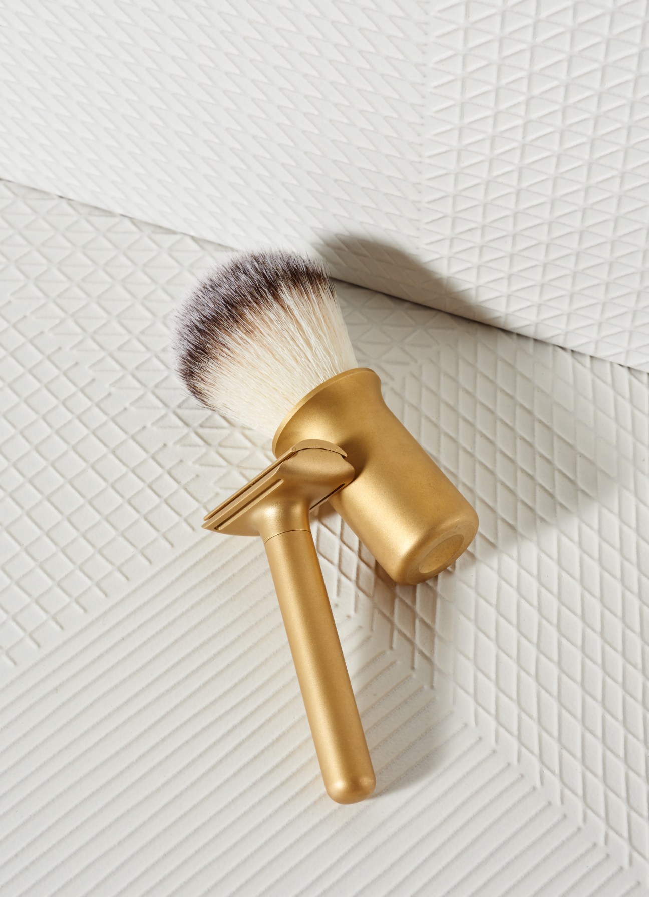 Gold-handled razor and brush with soft bristles against white textured tile background.